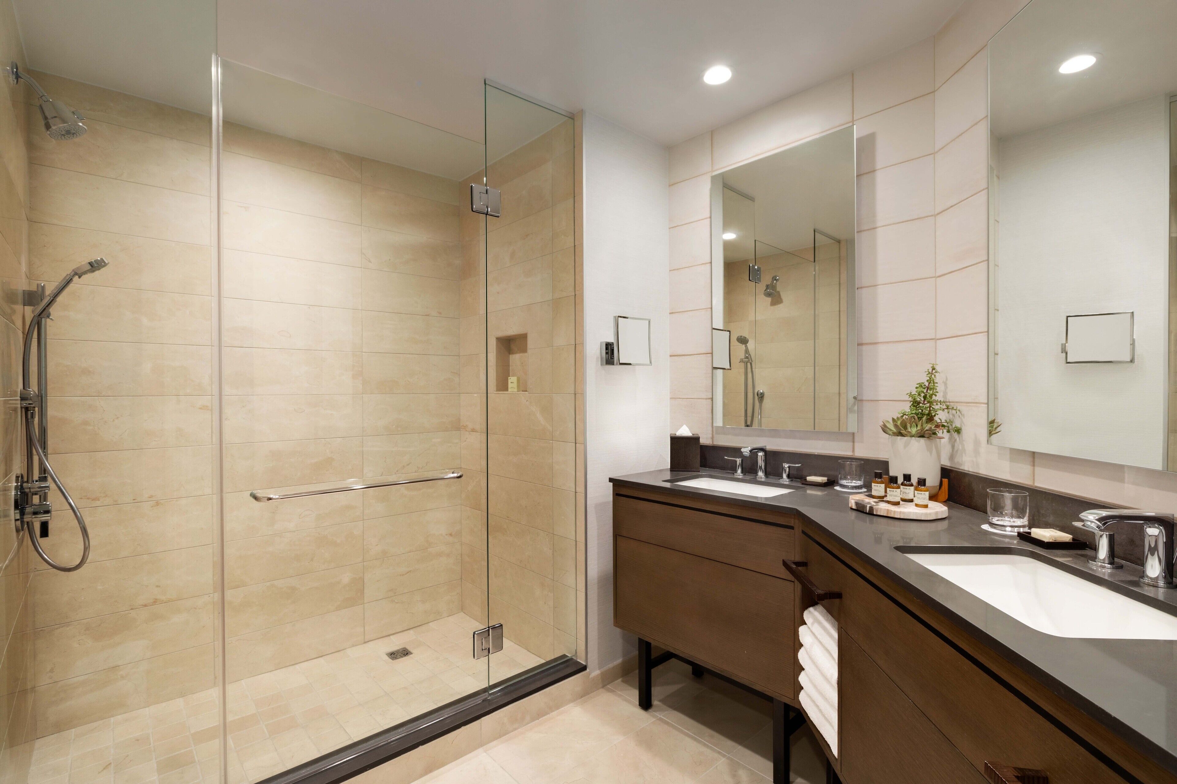 A modern bathroom with a glass-enclosed shower, two sinks, a large mirror, and a small plant on the vanity.