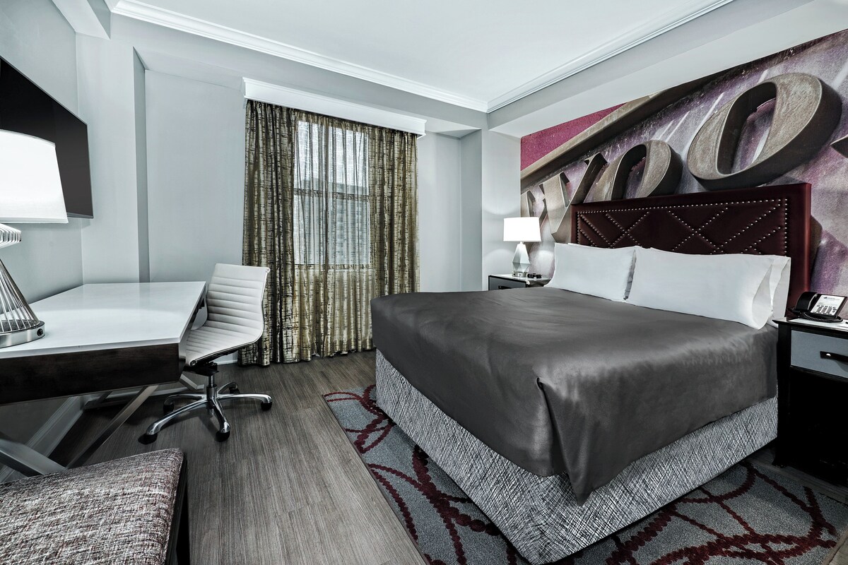 A hotel room with a large bed, a desk, a chair, a nightstand, and a large window with curtains.