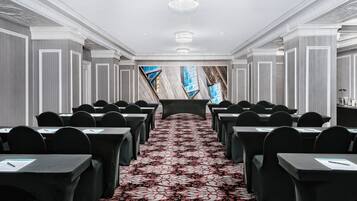 A conference room with rows of tables and chairs, a patterned carpet, and a large window with a view of the sky.