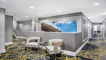 A hotel corridor with a patterned carpet, two chairs, a small table, and a large window with a view of a building.