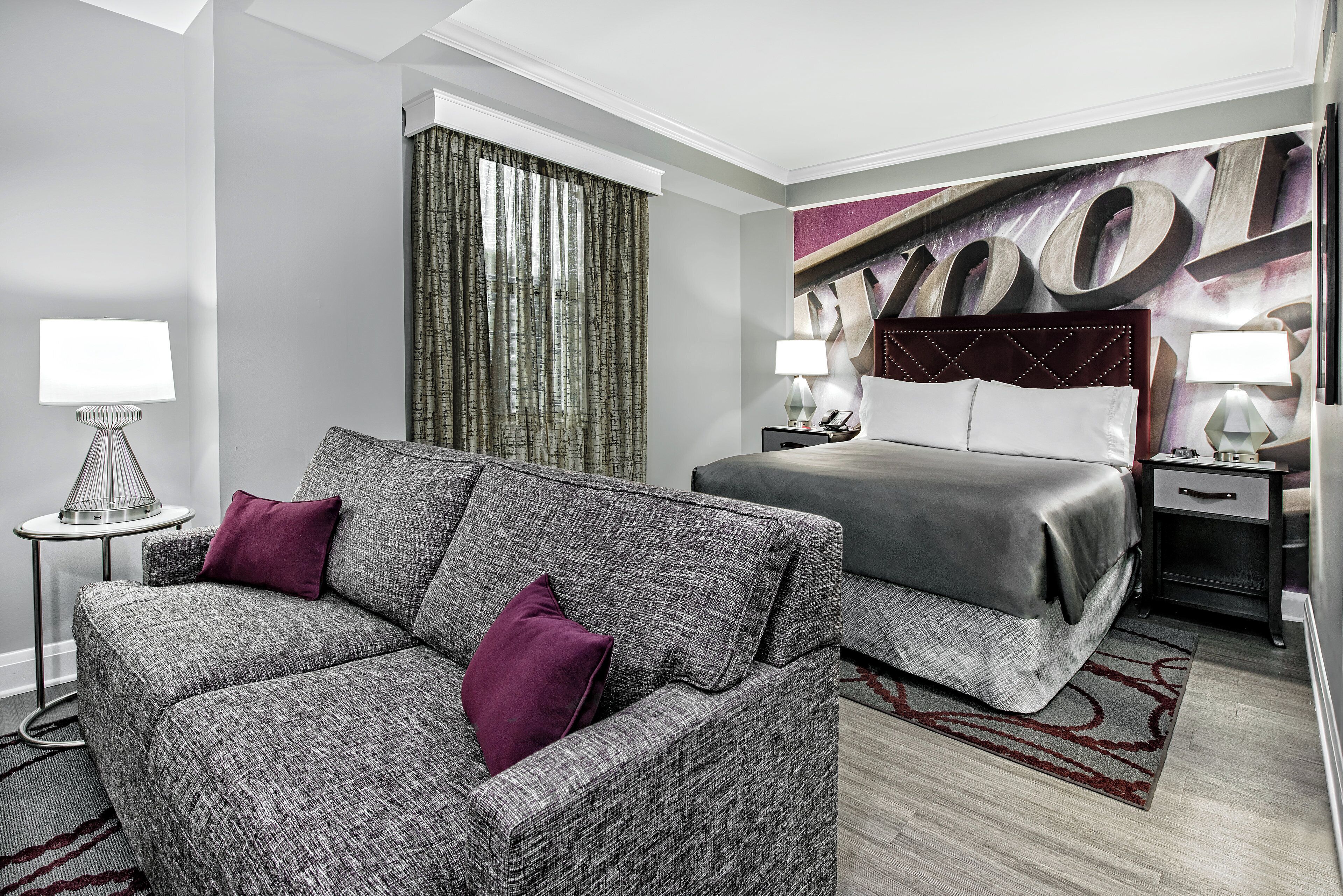 Habitación de hotel con un sofá gris, una cama con cubrecama gris, un cabecero con un letrero grande que dice 'WOOD', una mesita de noche con una lámpara y una cortina.