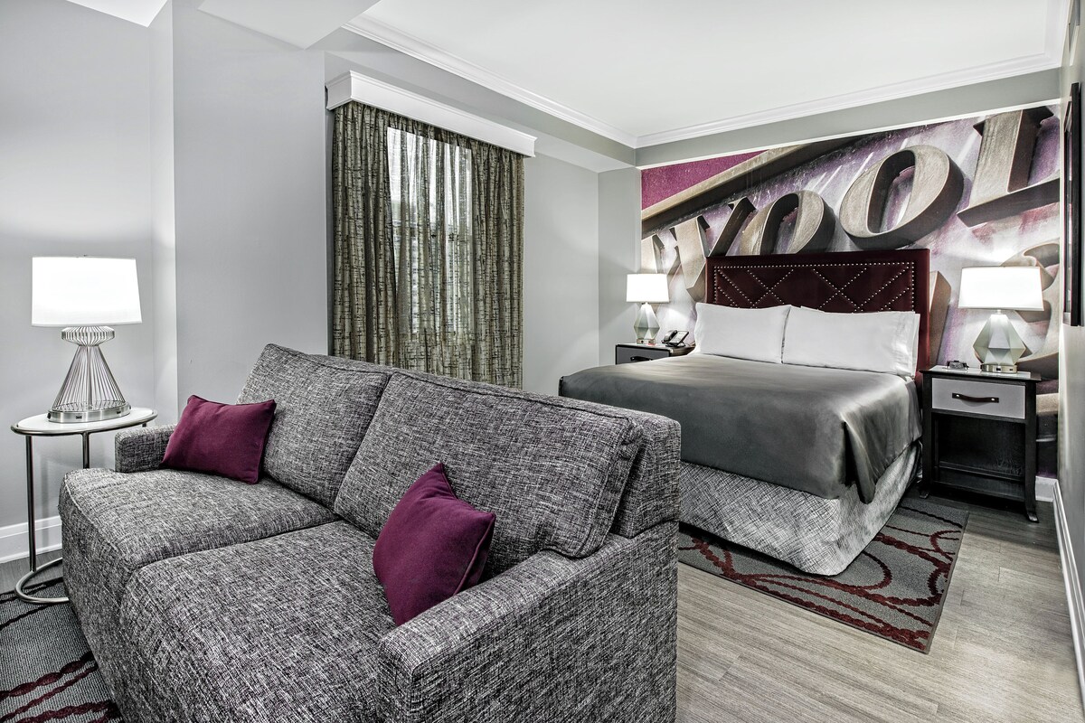A hotel room with a grey sofa, a bed with a grey bedspread, a headboard with a large 'WOOD' sign, a side table with a lamp, and a curtain.