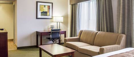 A hotel room with a bed, a sofa, a desk with a chair, a lamp, and a framed picture on the wall.