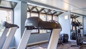 Ett gym med löpband och annan träningsutrustning, stora fönster och utsikt över byggnaderna utanför.