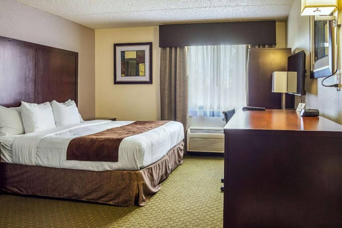 A hotel room with a bed, a desk, a chair, a lamp, a picture on the wall, and a window with curtains.