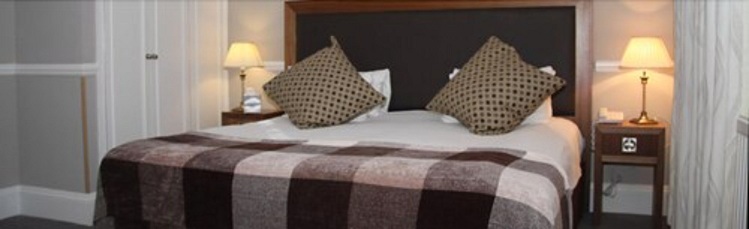 A hotel room with a large bed, two pillows, a checkered blanket, and two bedside lamps.