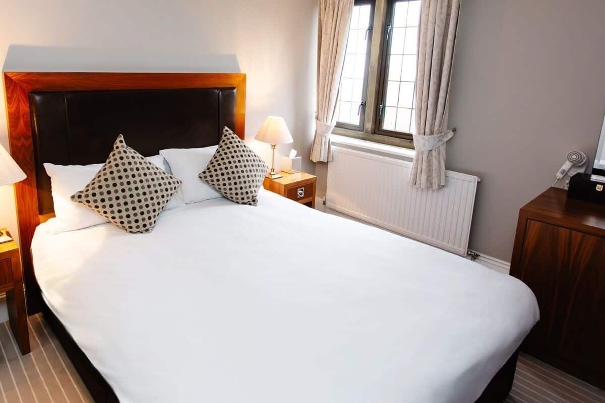A hotel room with a large bed, two bedside tables with lamps, a window with curtains, and a radiator.