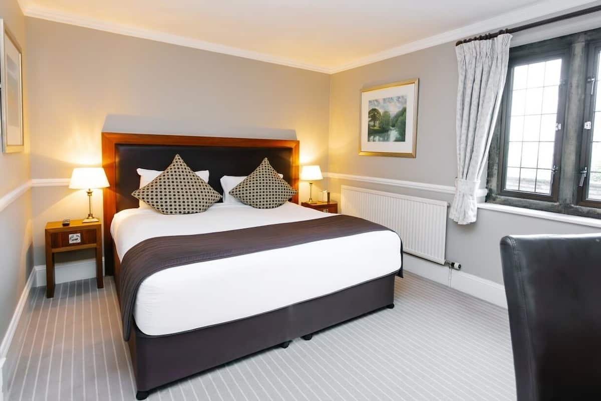 A hotel room with a large bed, two bedside tables with lamps, a radiator, a window with curtains, and a framed picture on the wall.