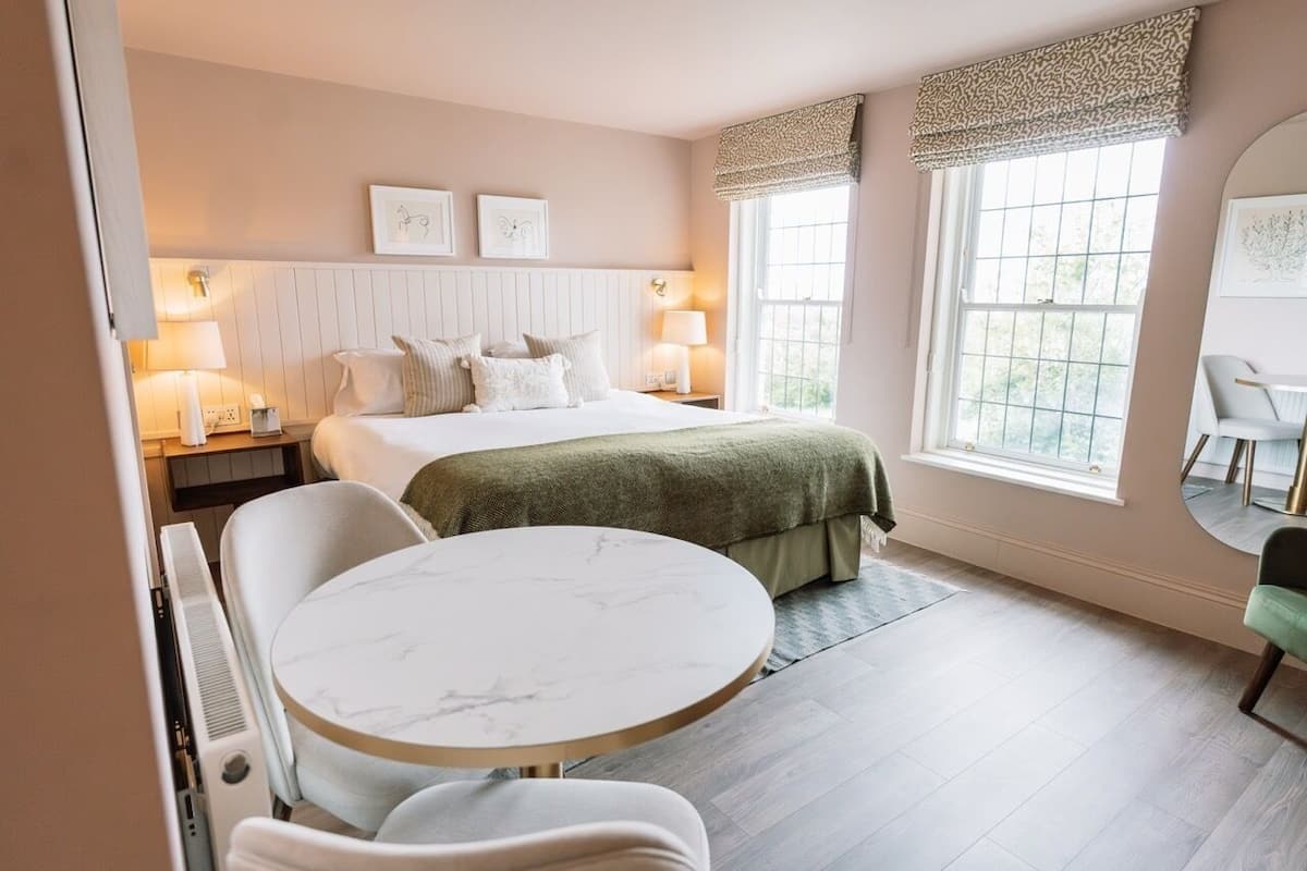 A hotel room with a bed, bedside tables, a round table, chairs, and large windows.