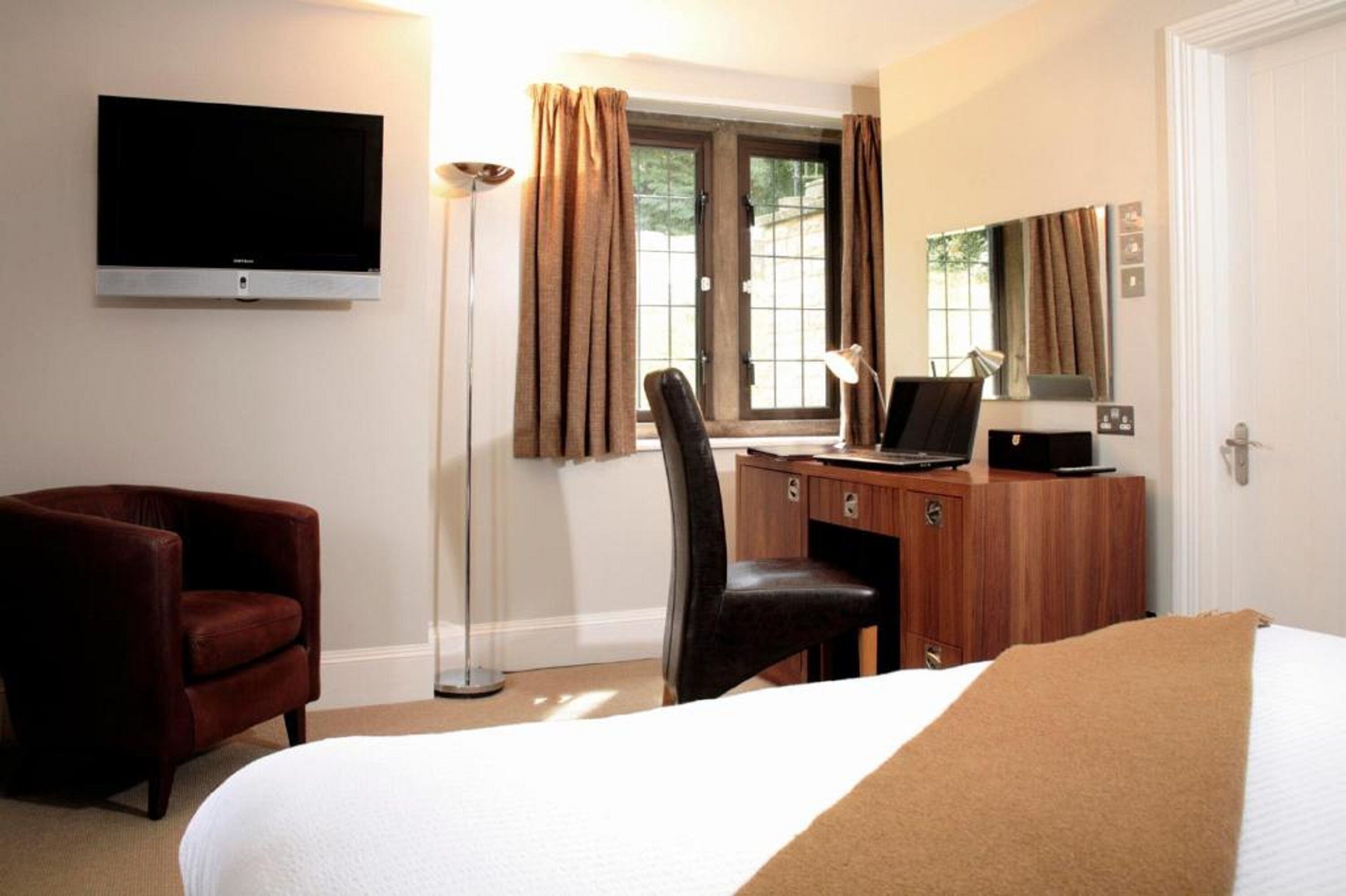 Quarto de hotel com cama, escrivaninha com computador, cadeira, televisão fixada na parede e janela com cortinas.