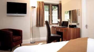 Quarto de hotel com cama, escrivaninha com computador, cadeira, televisão fixada na parede e janela com cortinas.