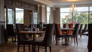 A restaurant with wooden flooring, dark upholstered chairs, and a chandelier. Tables are set with glasses and cutlery. - Hatton Court (Gloucester)