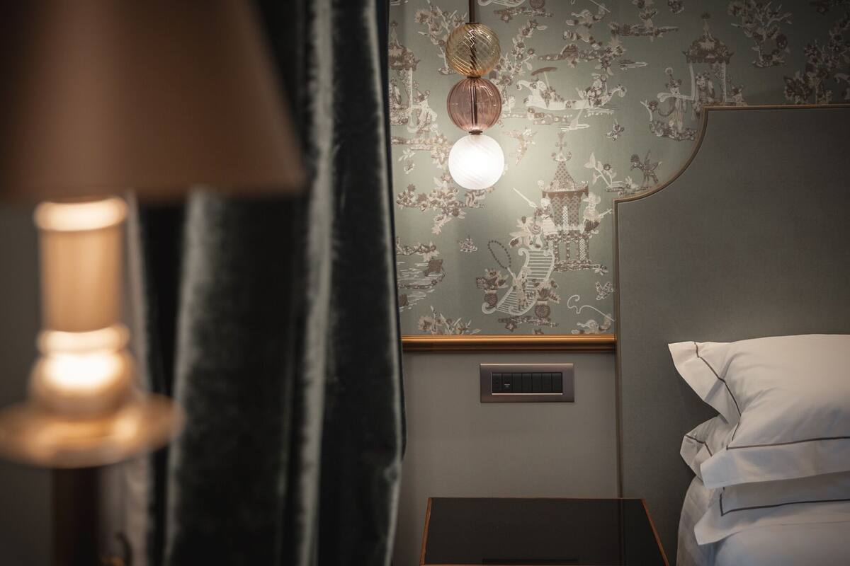 A hotel room with a bed, a nightstand, a wall-mounted lamp, and a decorative wall with a floral pattern.