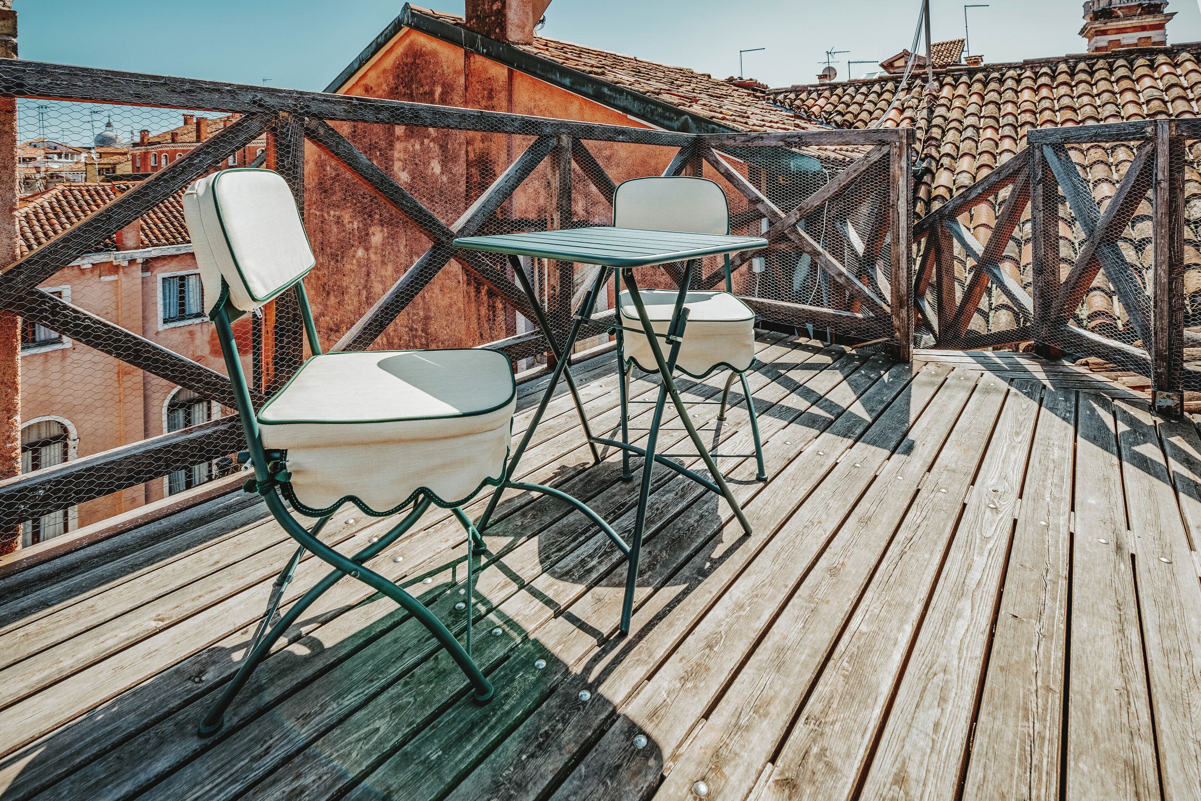 A wooden deck with metal chairs and a table, overlooking a cityscape with terracotta roofs.