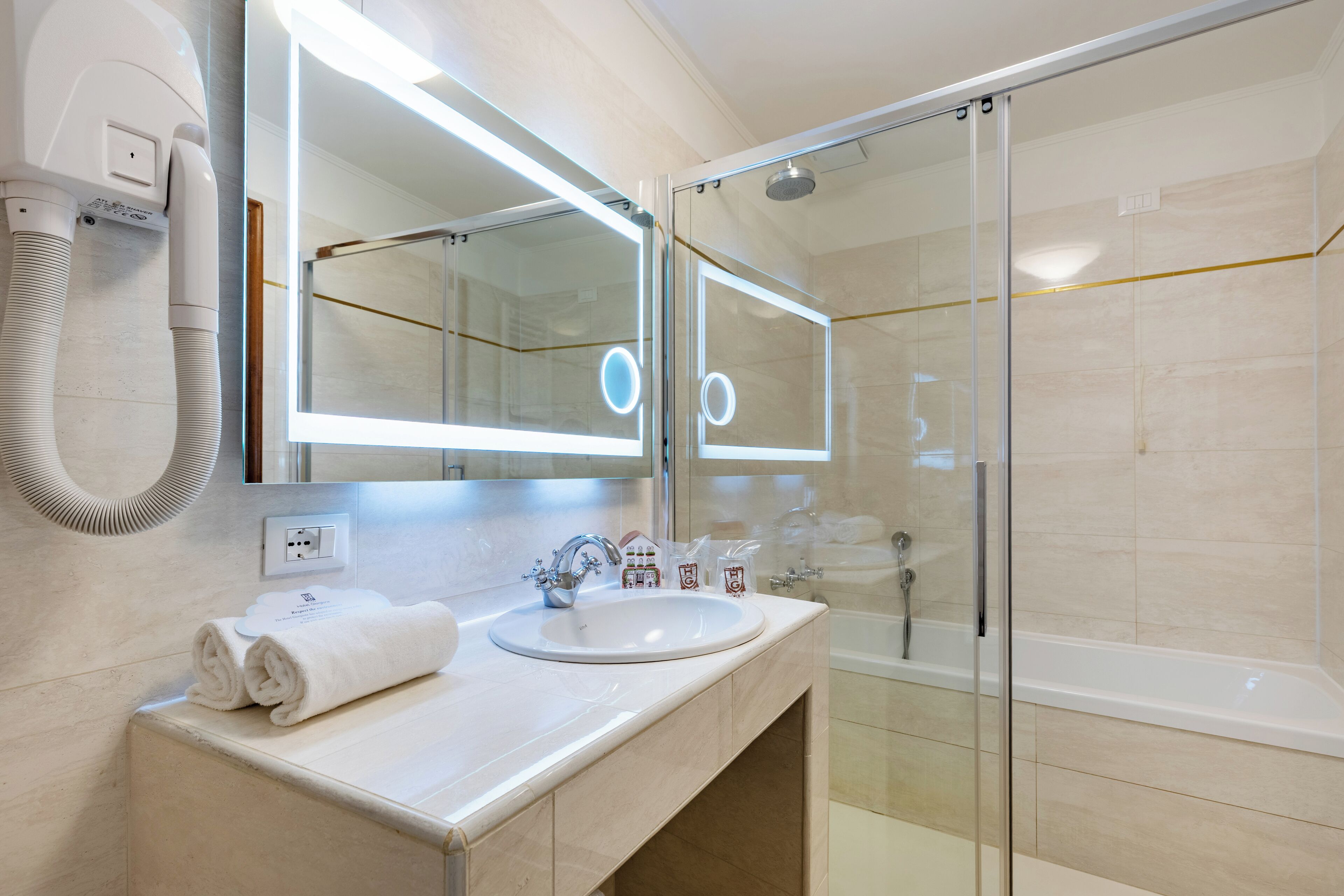 Un baño moderno con ducha envuelta en vidrio, un lavabo blanco y un espejo grande con iluminación integrada.