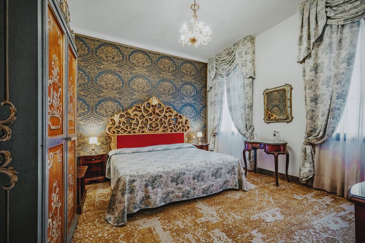 A room with a large bed, ornate headboard, patterned wallpaper, and a chandelier.