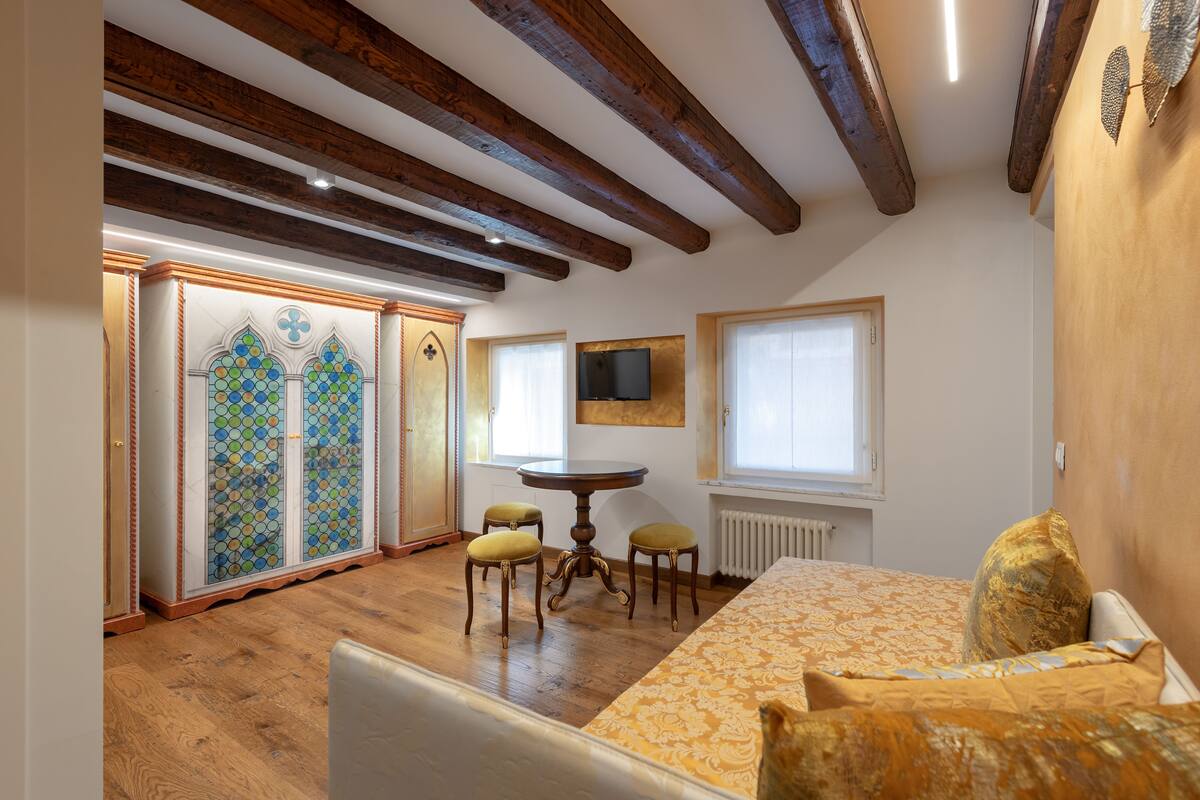 A bedroom with wooden beams, a bed with patterned bedding, a small table, two chairs, and a decorative cabinet with glass doors.