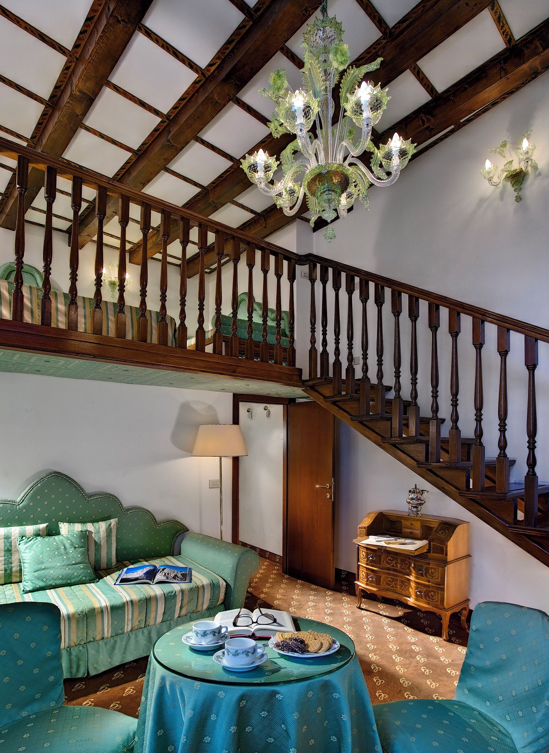 A room with a wooden staircase, a green sofa set, a table with tea cups and biscuits, and a chandelier.