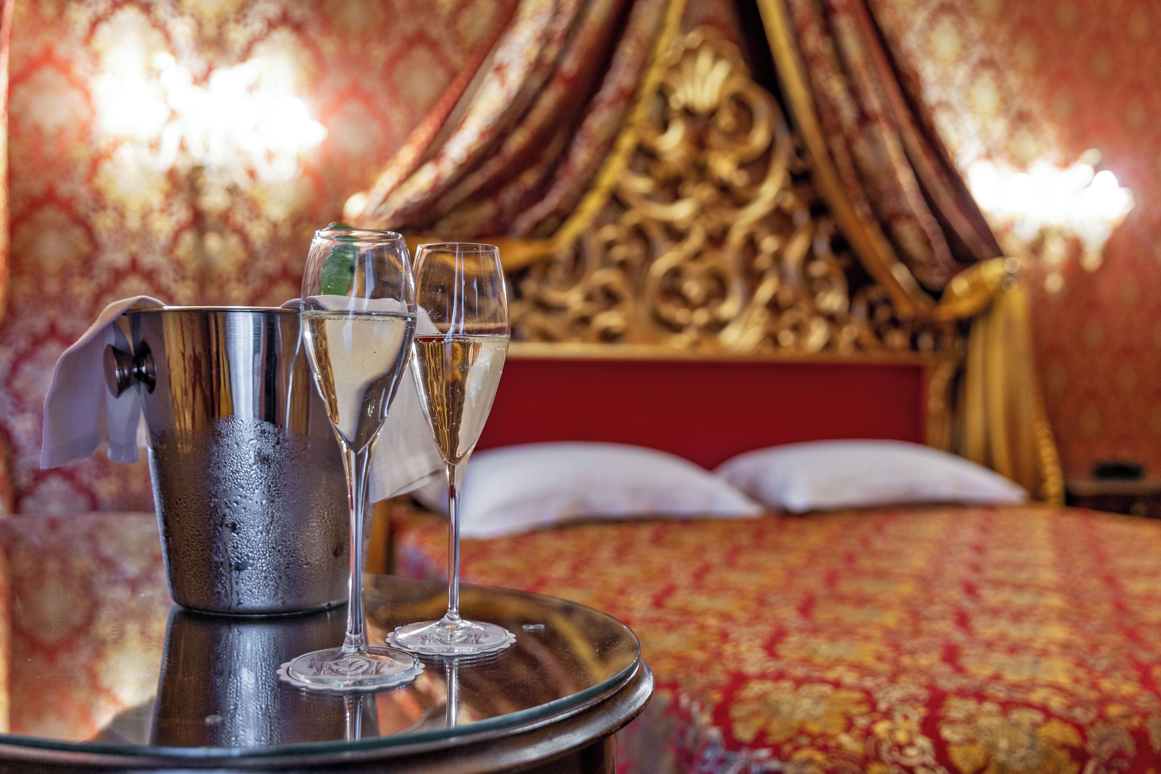 A bed with a red and gold patterned bedspread, a metal tray with two glasses of champagne, and a decorative metal pitcher.