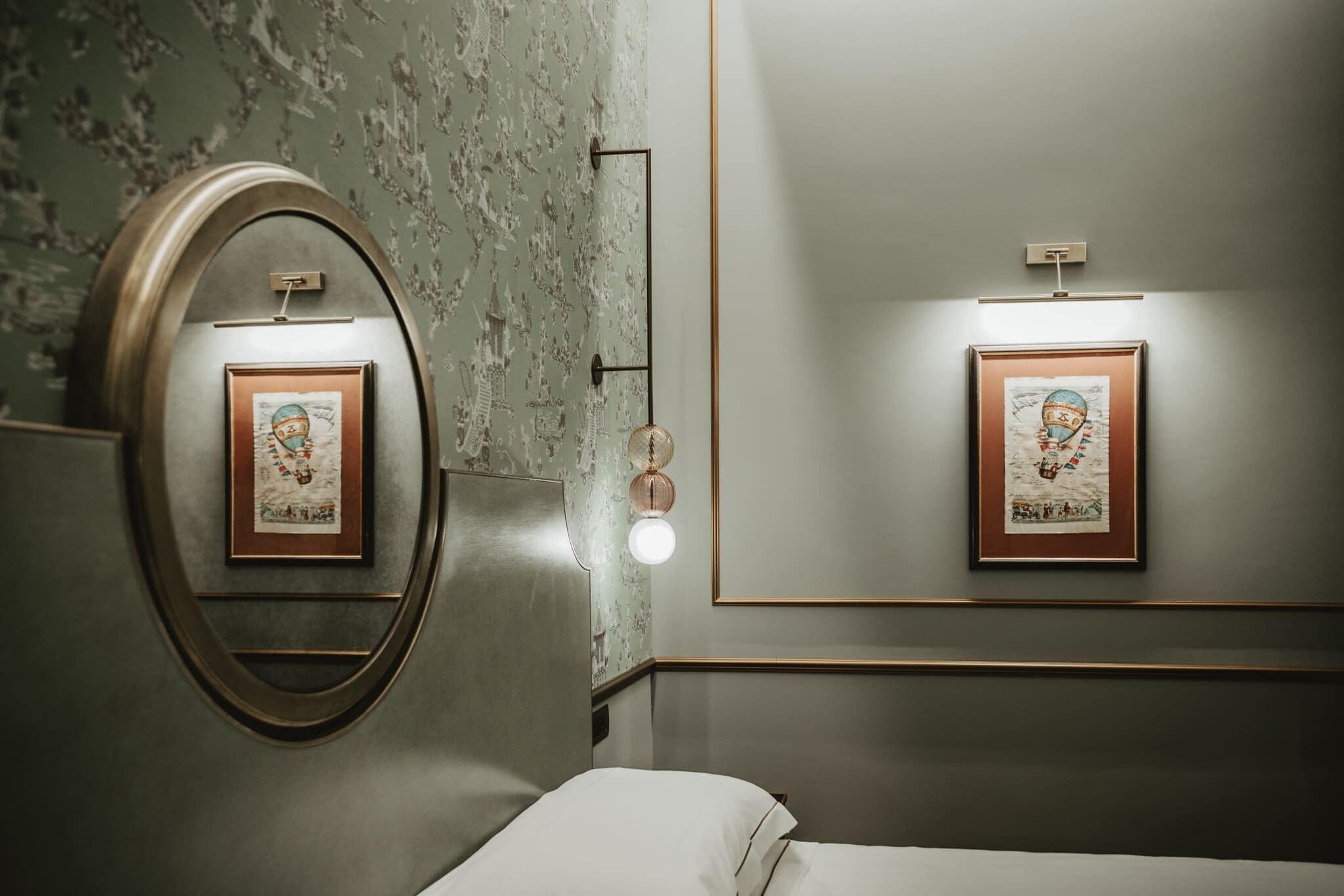 A hotel room with a large mirror, two framed artworks on the wall, a bedside lamp, and a neatly made bed.