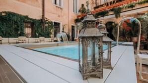 Uma área na piscina com uma mesa branca, um candeeiro decorativo e um edifício com janelas em arco e vegetação.