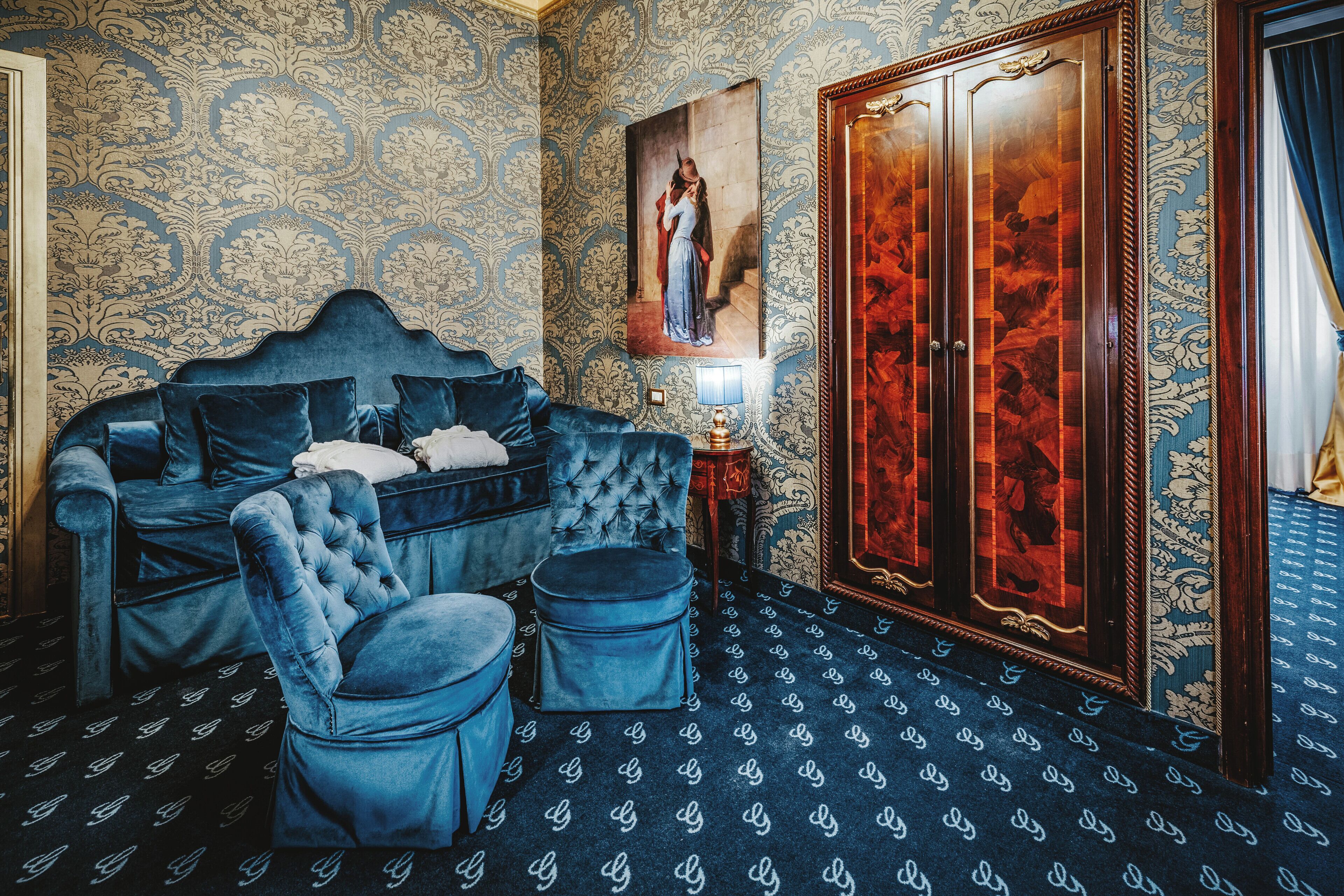 A room with a tufted blue sofa, matching ottoman, and a wooden wardrobe with a mirror.