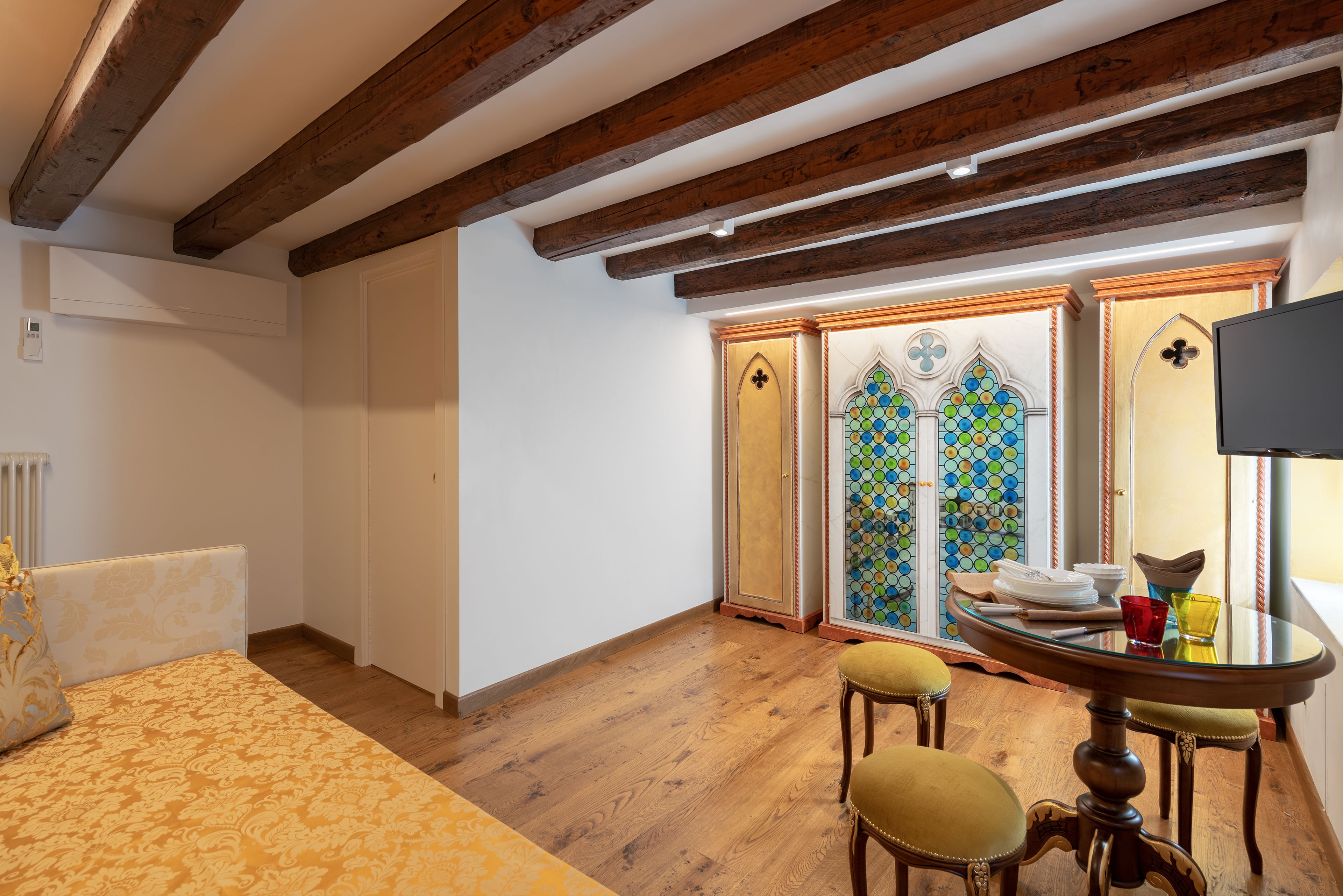 A room with wooden beams, a bed with a patterned bedspread, a dining table with chairs, and a stained glass window.