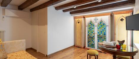A room with wooden beams, a bed with a patterned bedspread, a dining table with chairs, and a stained glass window.