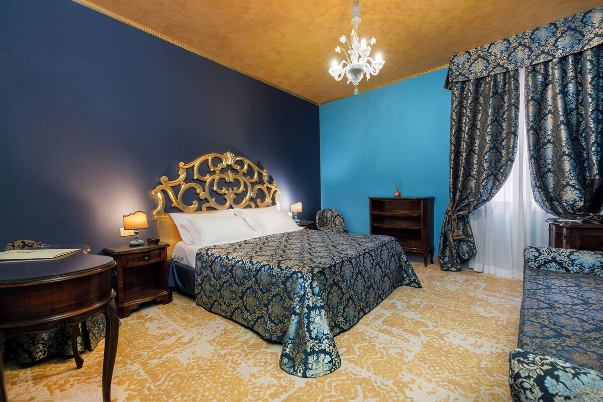 A bedroom with a large bed, a nightstand, a chair, a desk, and a chandelier.