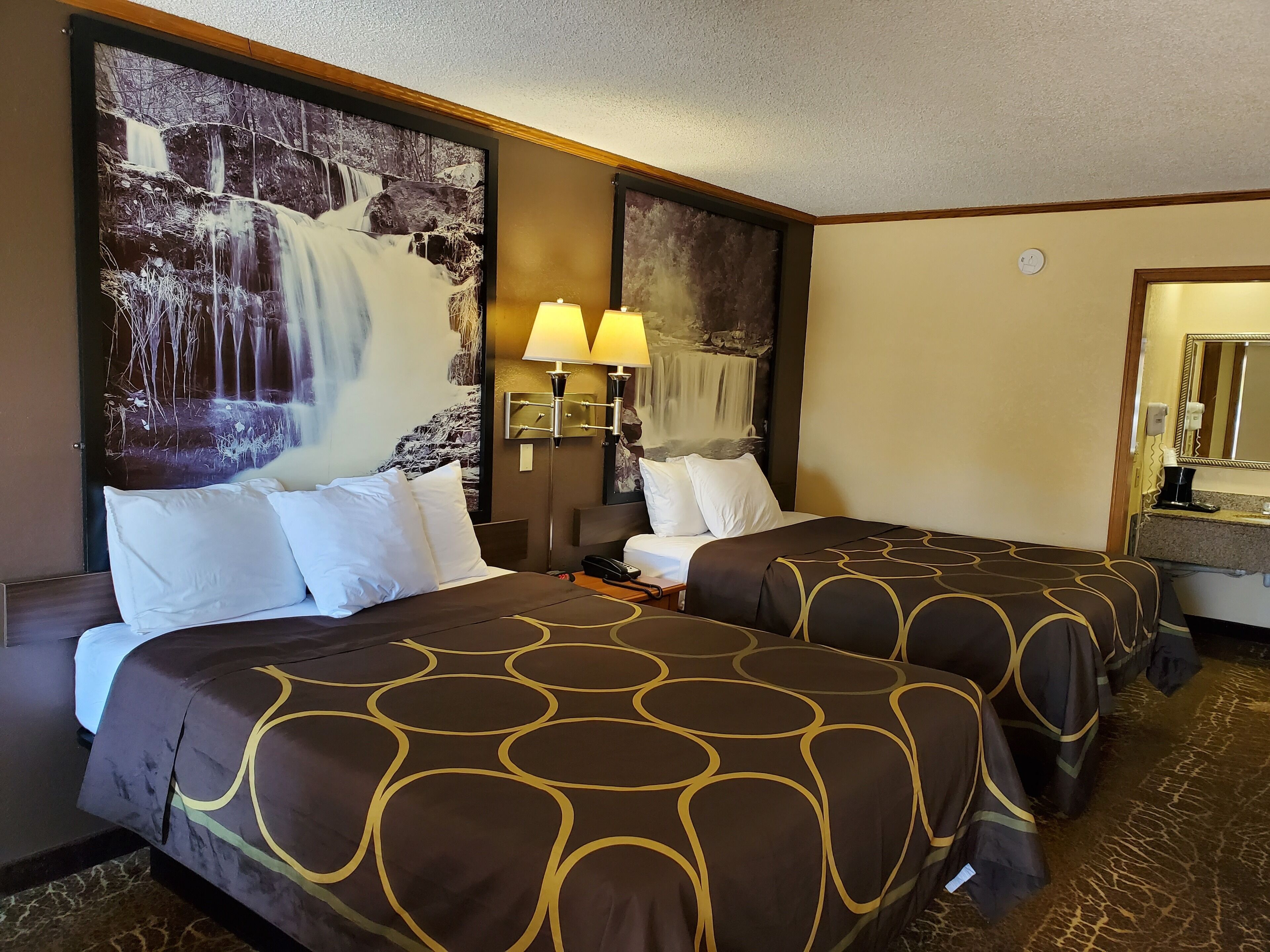 Habitación de hotel con dos camas, un mural grande de una cascada en la pared, una mesita de noche con lámpara y un espejo sobre el lavamanos.