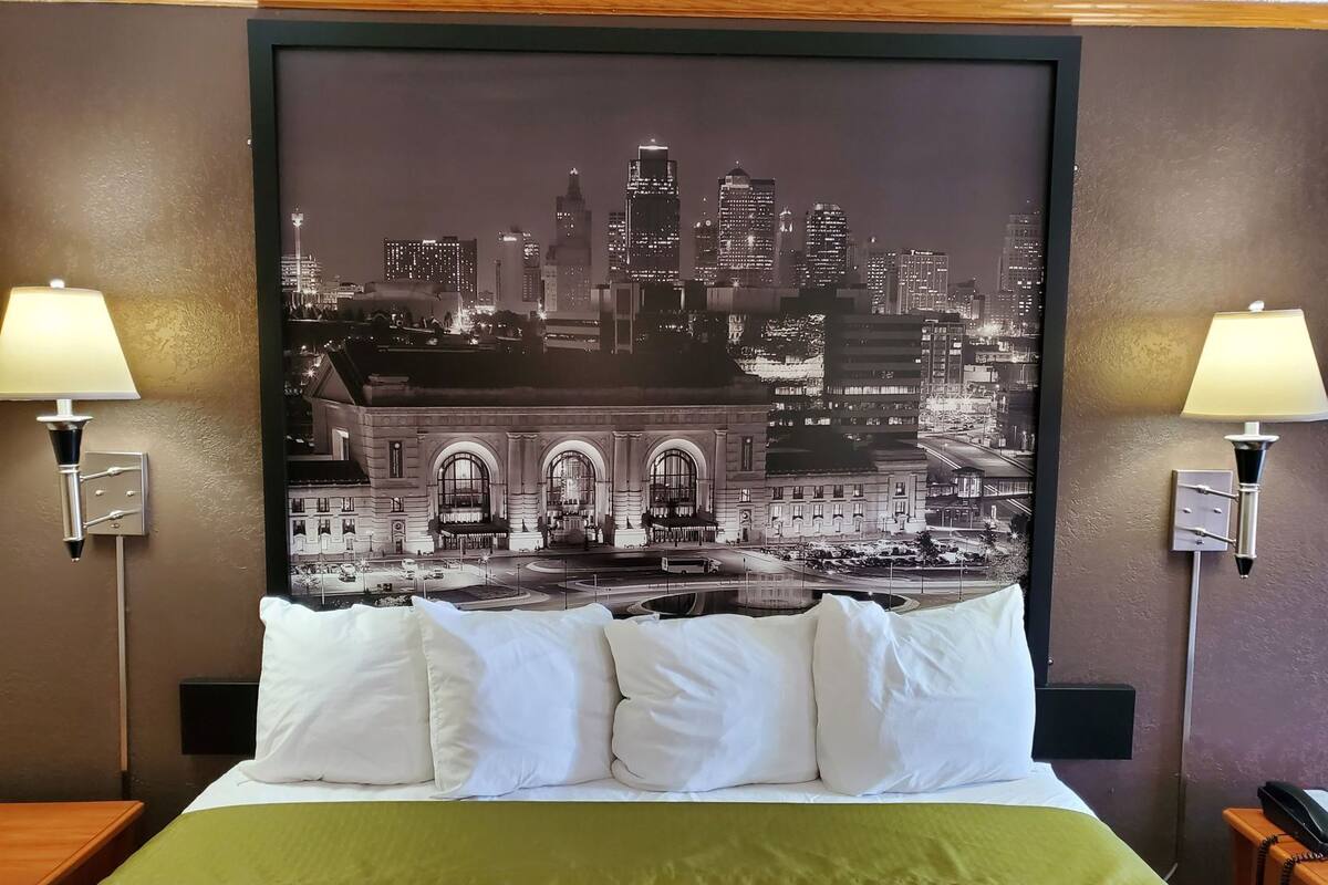 Una camera d'albergo con un grande quadro raffigurante un panorama cittadino, un letto con cuscini bianchi, una coperta verde e due lampade da comodino.