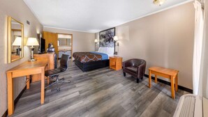 A hotel room with a bed, a desk, a chair, a small table, and a lamp.
