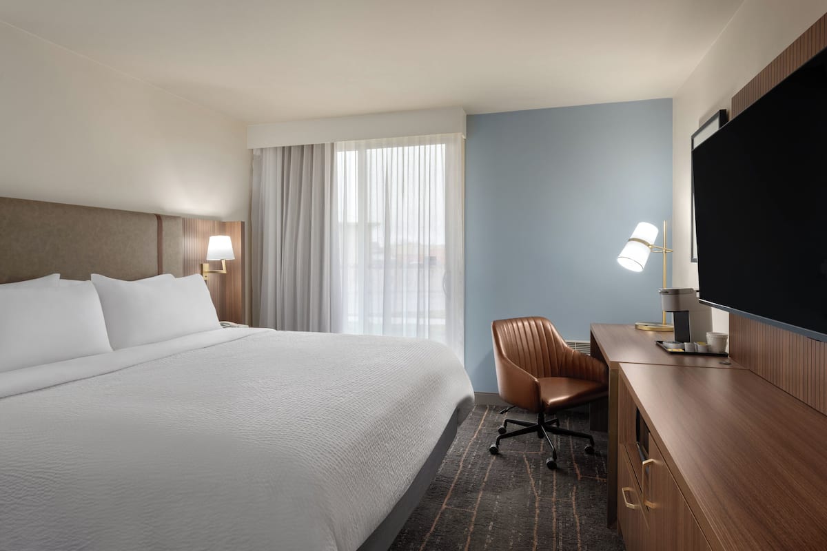 A hotel room with a large bed, a desk with a chair, a flat-screen TV, a lamp, and a window with sheer curtains.