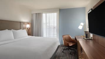 A hotel room with a large bed, a desk with a chair, a flat-screen TV, and a window with curtains.
