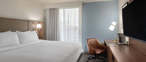 A hotel room with a large bed, a desk with a chair, a flat-screen TV, and a window with curtains.