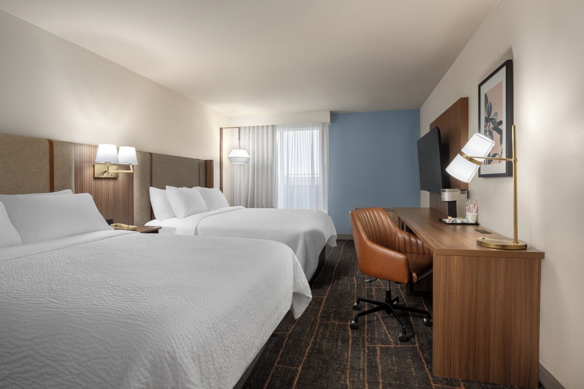 A hotel room with two beds, a desk, a chair, a lamp, and a window with curtains.