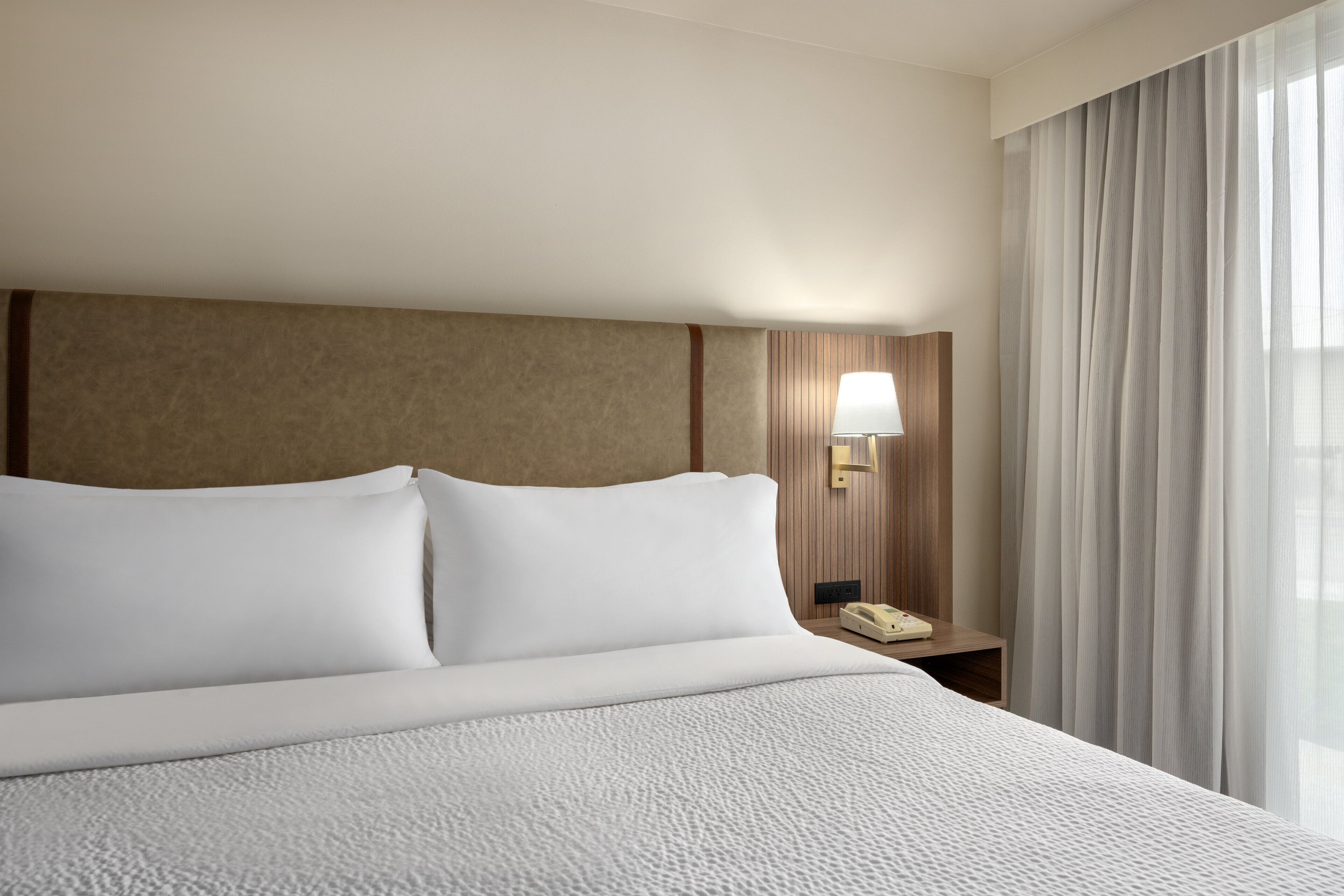 A hotel room with a large bed, a bedside lamp, a phone, and sheer curtains.