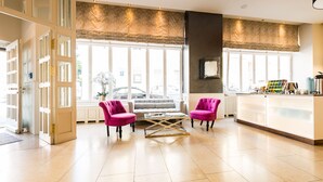 A hotel lobby with a reception desk, two pink armchairs, a sofa, and large windows with a view of the street. - The Heidelberg Exzellenz Hotel (Heidelberg)
