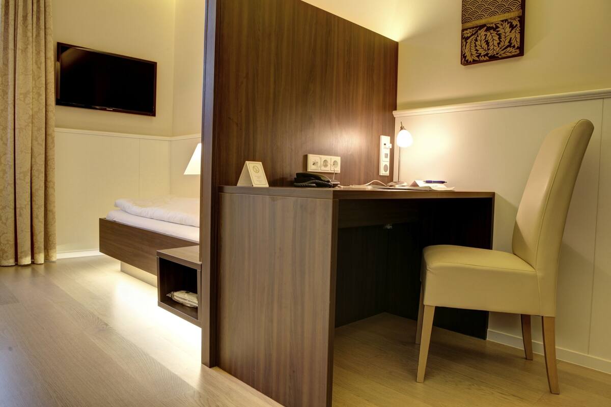 A compact hotel room with a bed, a desk with a chair, a television, and a lamp.