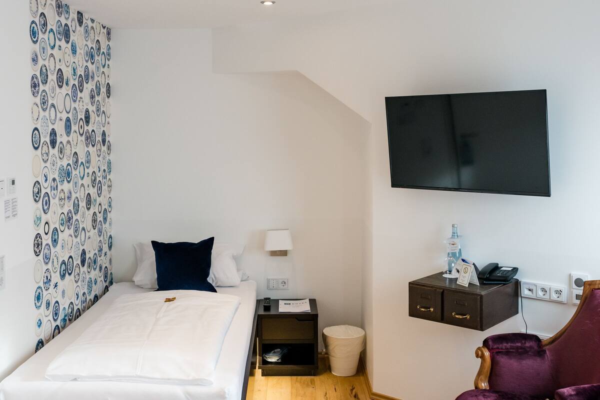 A modern hotel room with a wooden floor, a bed with white linens, a purple armchair, a TV mounted on the wall, and a patterned curtain.