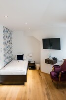 Een moderne hotelkamer met een houten vloer, een bed met witte lakens, een paarse fauteuil, een aan de muur bevestigde televisie en een gordijn met dessin.