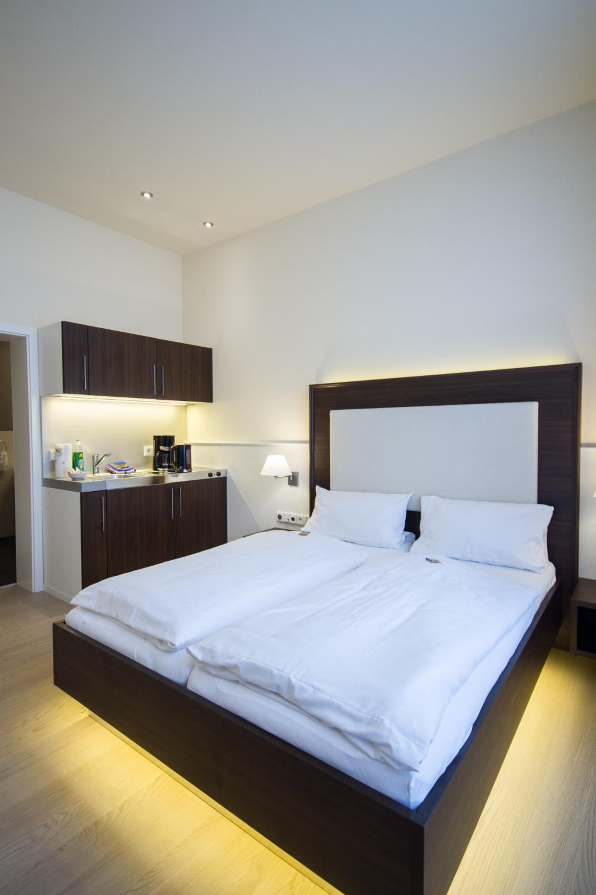 Een moderne hotelkamer met een groot bed, een kleine kitchenette en een nachtkastje met een lamp.