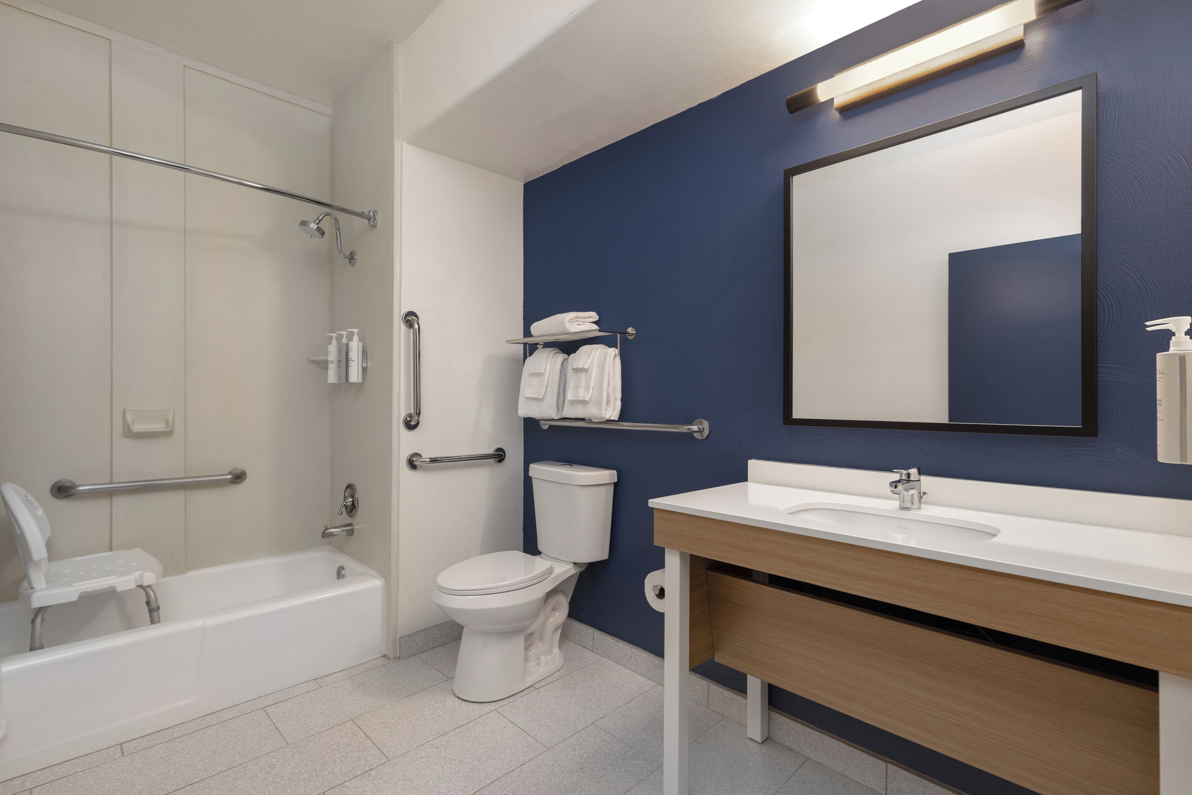A modern bathroom with a bathtub, toilet, and sink, equipped with grab bars and a mirror.