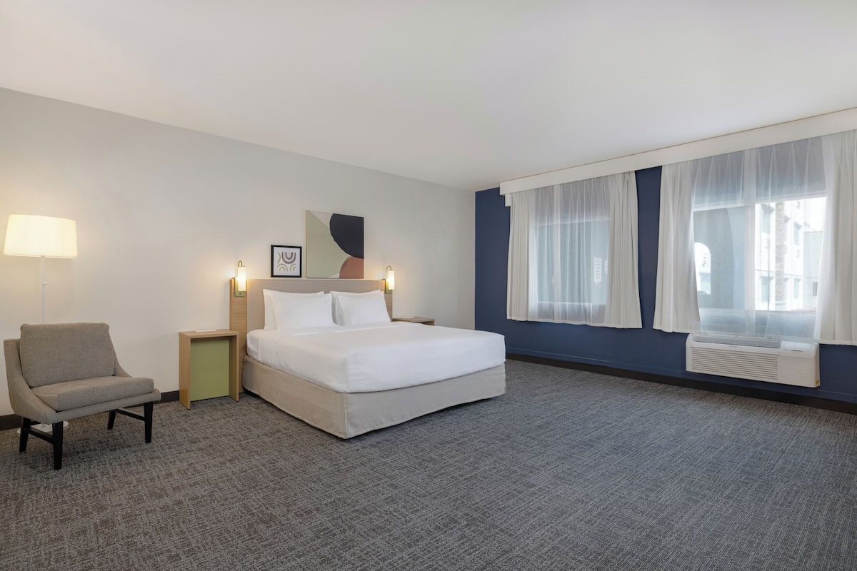 Hotellihuone, jossa on sänky, tuoli, yöpöytä, lamppu, taulu ja ikkuna, jossa on verhot.