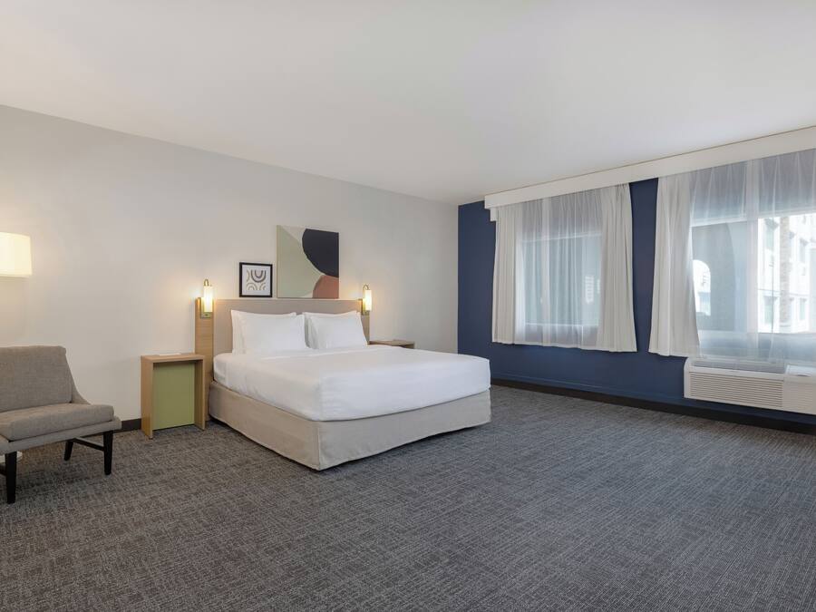 Habitación de hotel con cama, una silla, mesita de noche, lámpara, un cuadro y ventana con cortinas.