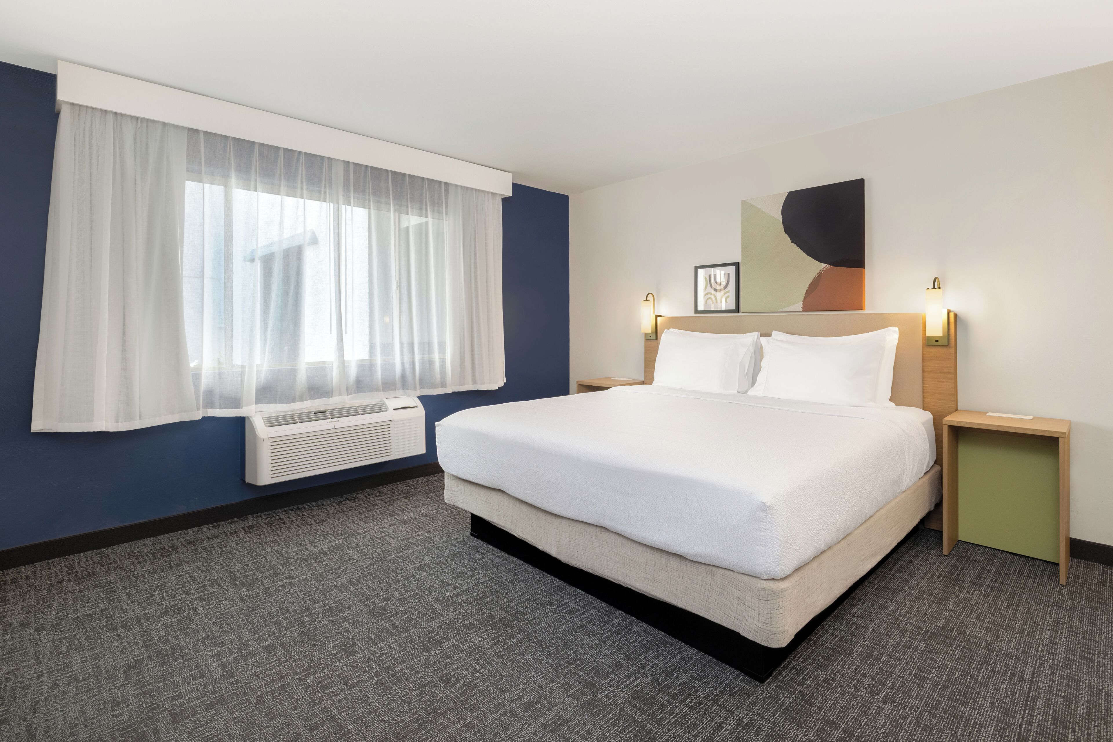 A hotel room with a large bed, a window with sheer curtains, a wall-mounted air conditioner, a bedside table with a lamp, and a framed picture on the wall.
