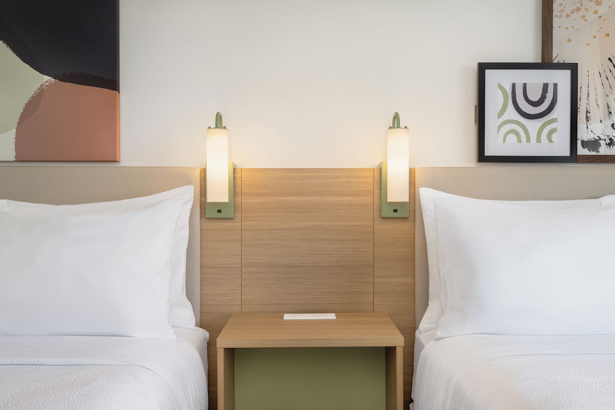 Hotellihuone, jossa on kaksi sänkyä, puinen pääty, yöpöytävalaisimet, pieni pöytä ja taideteoksia seinillä.