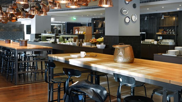 A modern restaurant with a long counter, multiple seating areas, and copper pendant lighting. - Scandic Park Helsinki (Helsinki)