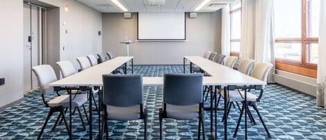 Une salle de conférence équipée d’une longue table, de chaises, d’un écran de projection et de grandes fenêtres.