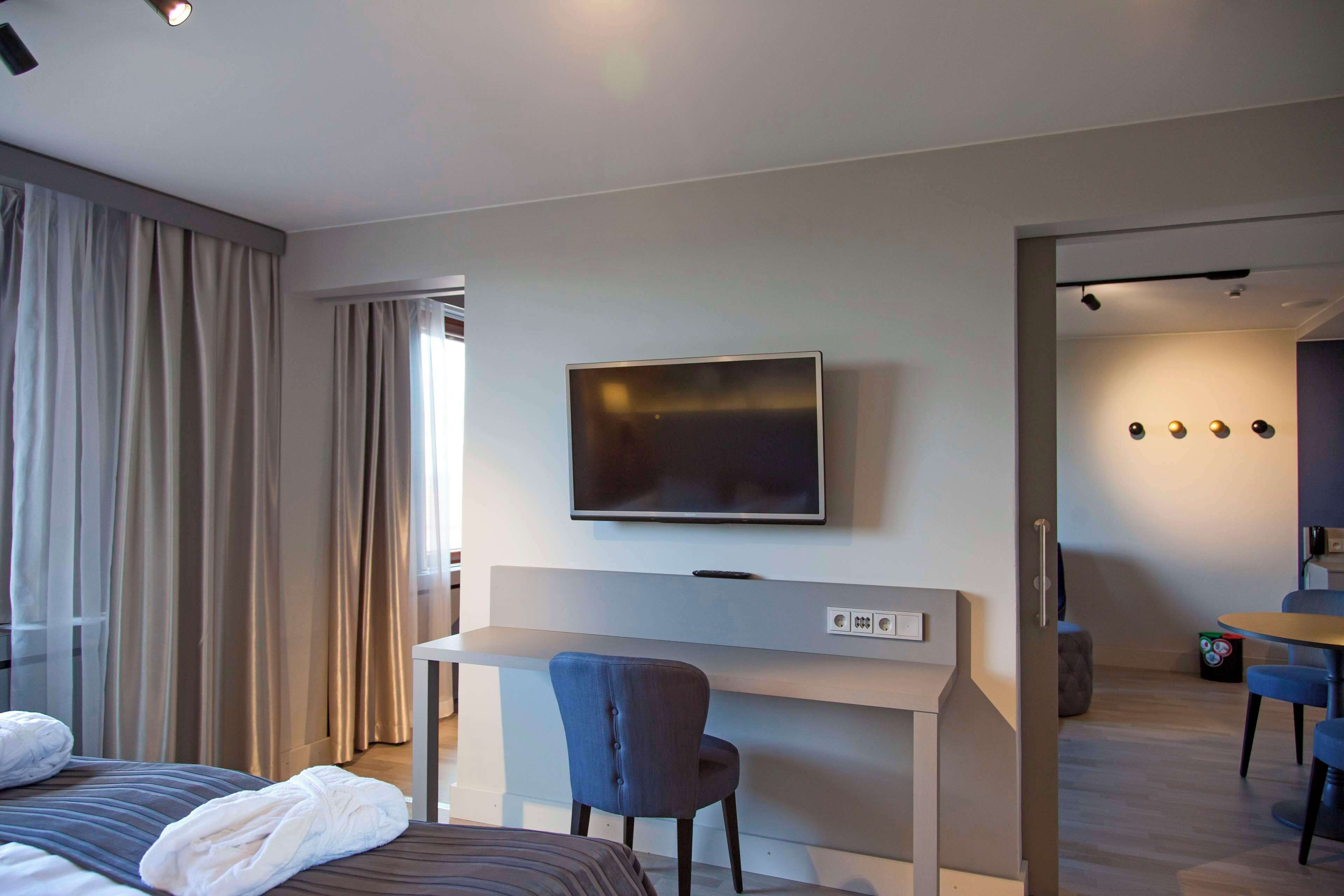 Una habitación de hotel moderna con cama, escritorio con silla, televisión y zona de comedor con sillas.